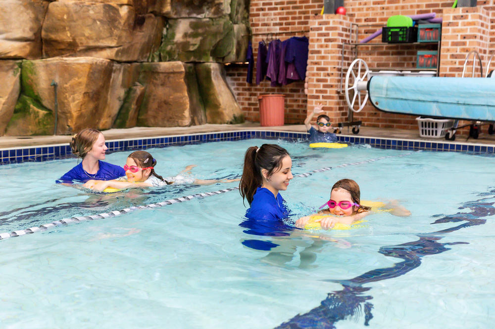 Two swim instructors in blue shirts teaching children in a pool. The children wear goggles and flotation devices.
