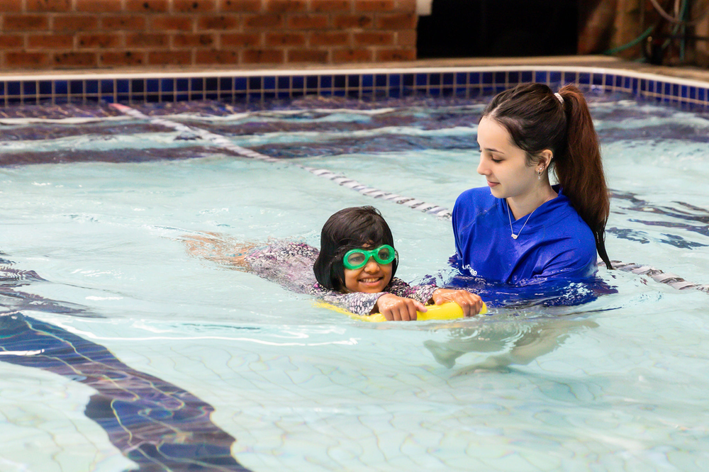 A swim instructor assists a smiling child wearing goggles who is using a yellow kickboard in an indoor swimming pool.
