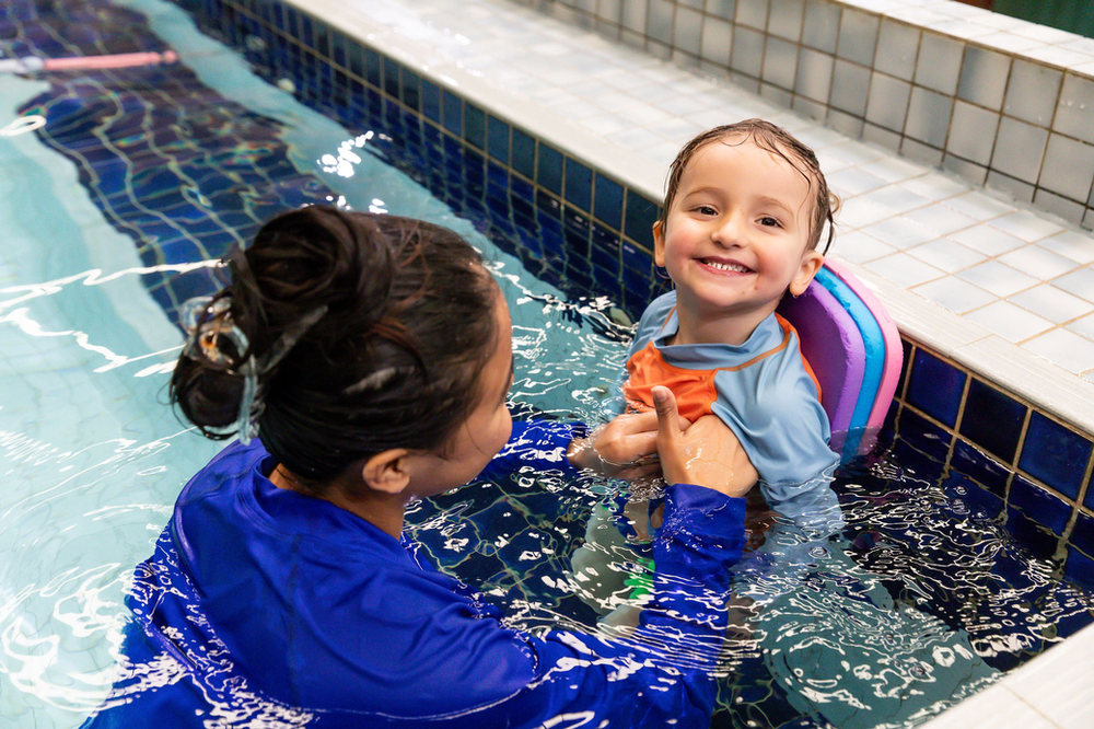 Child in pool with instructor, smiling. Instructor in blue shirt holding child. Pool has blue tiles.