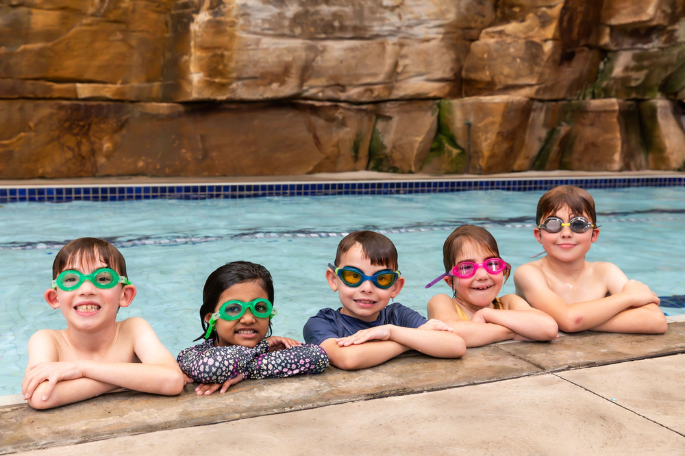 Five children wearing swim goggles pose with arms crossed at the edge of a swimming pool with a rock wall background.