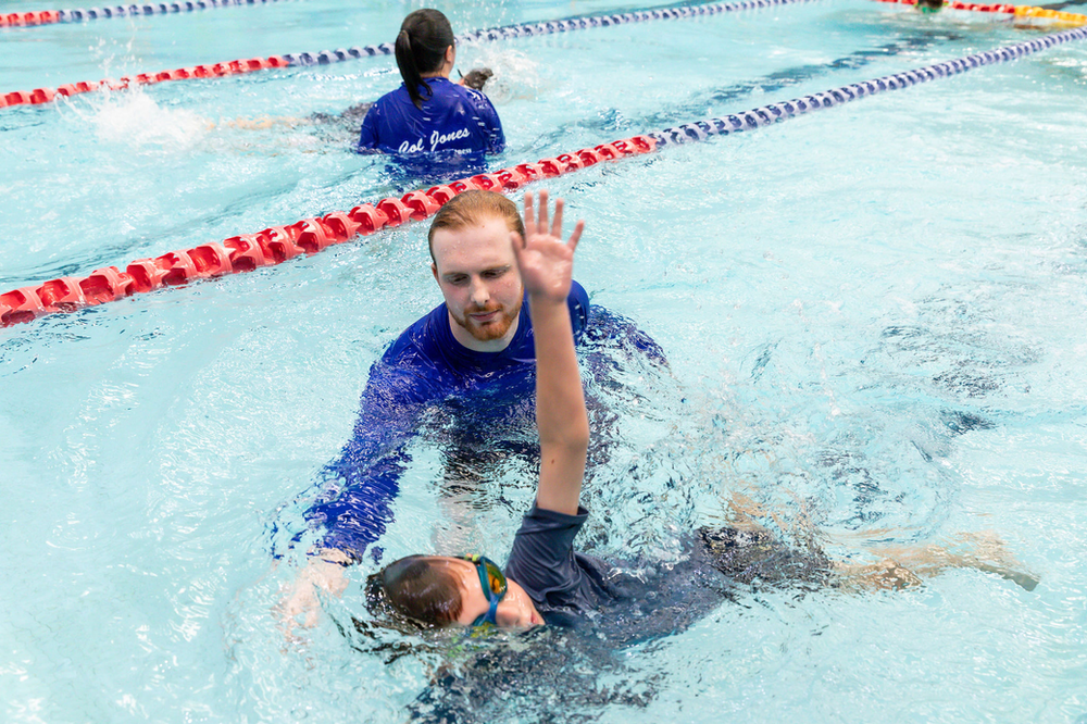 Swim instructor assisting a swimmer in a pool, with another swimmer in the background