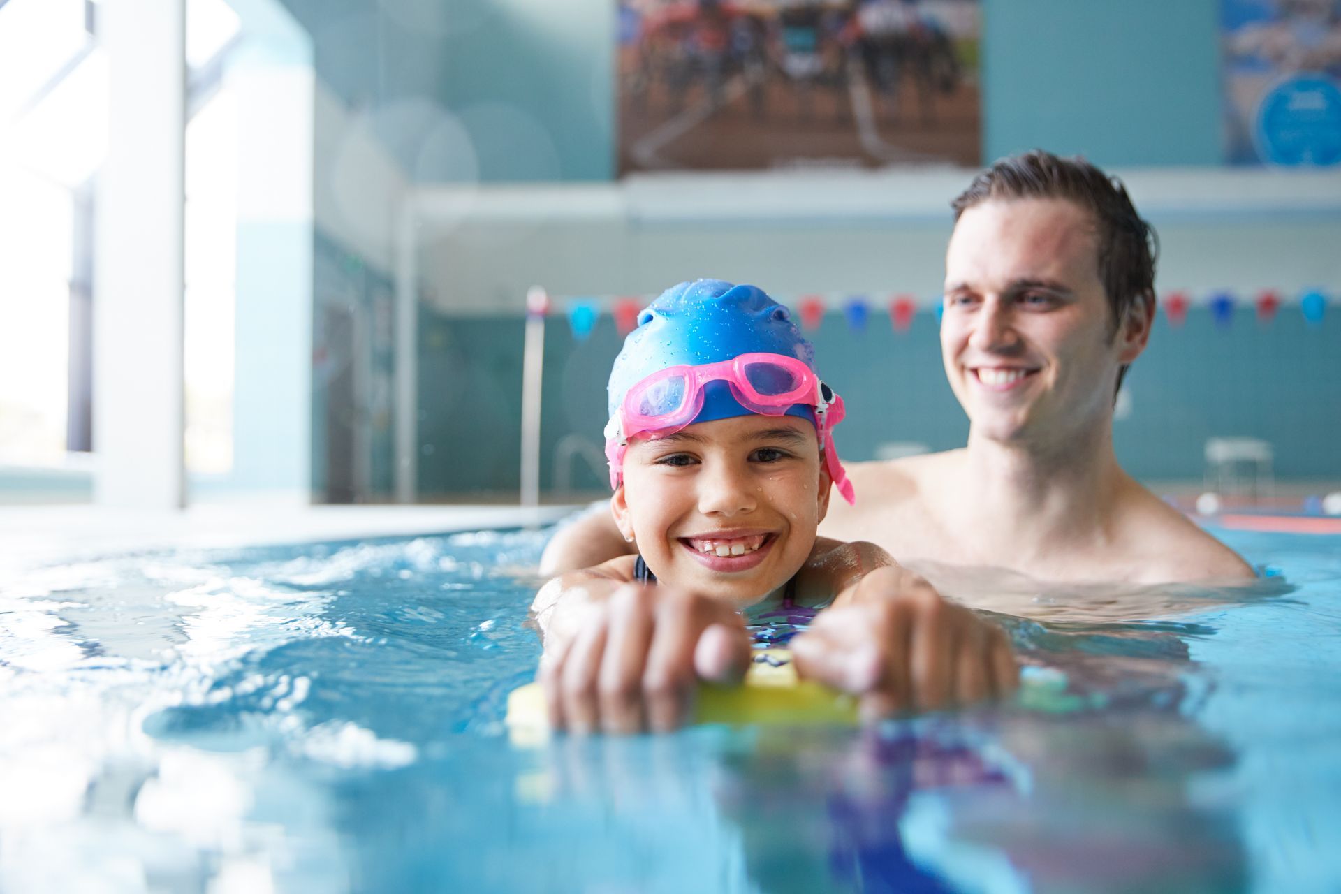 Girl in pool with swim instructor using a kickboard, smiling.