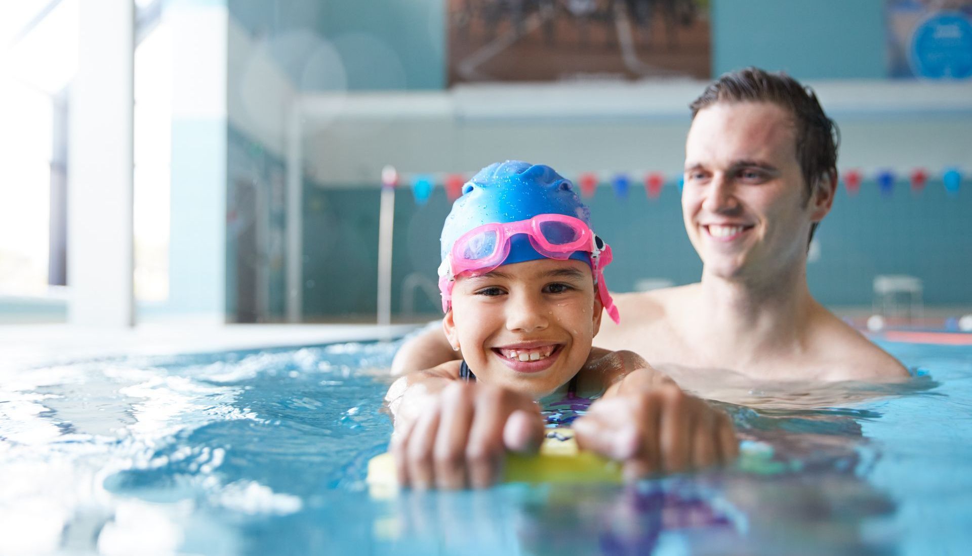 Girl with a swim cap and goggles smiles in pool, held by an instructor.