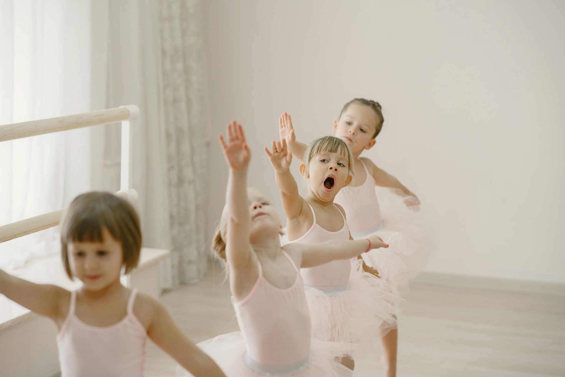 Four young children in pink leotards and tutus practice ballet movements in a bright, sunlit dance studio.