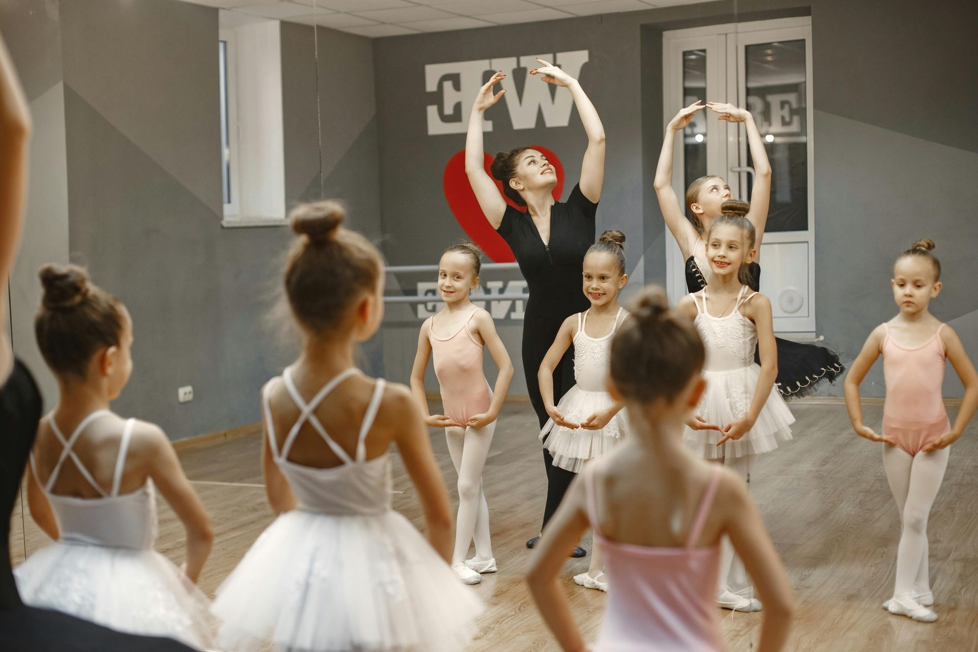 A dance instructor leads a group of children in ballet poses inside a studio with a heart wall decoration.