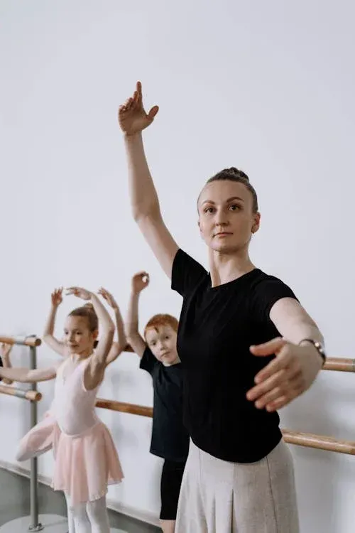 A ballet instructor leads a young student in a dance studio, both practicing arm positions in front of a barre.
