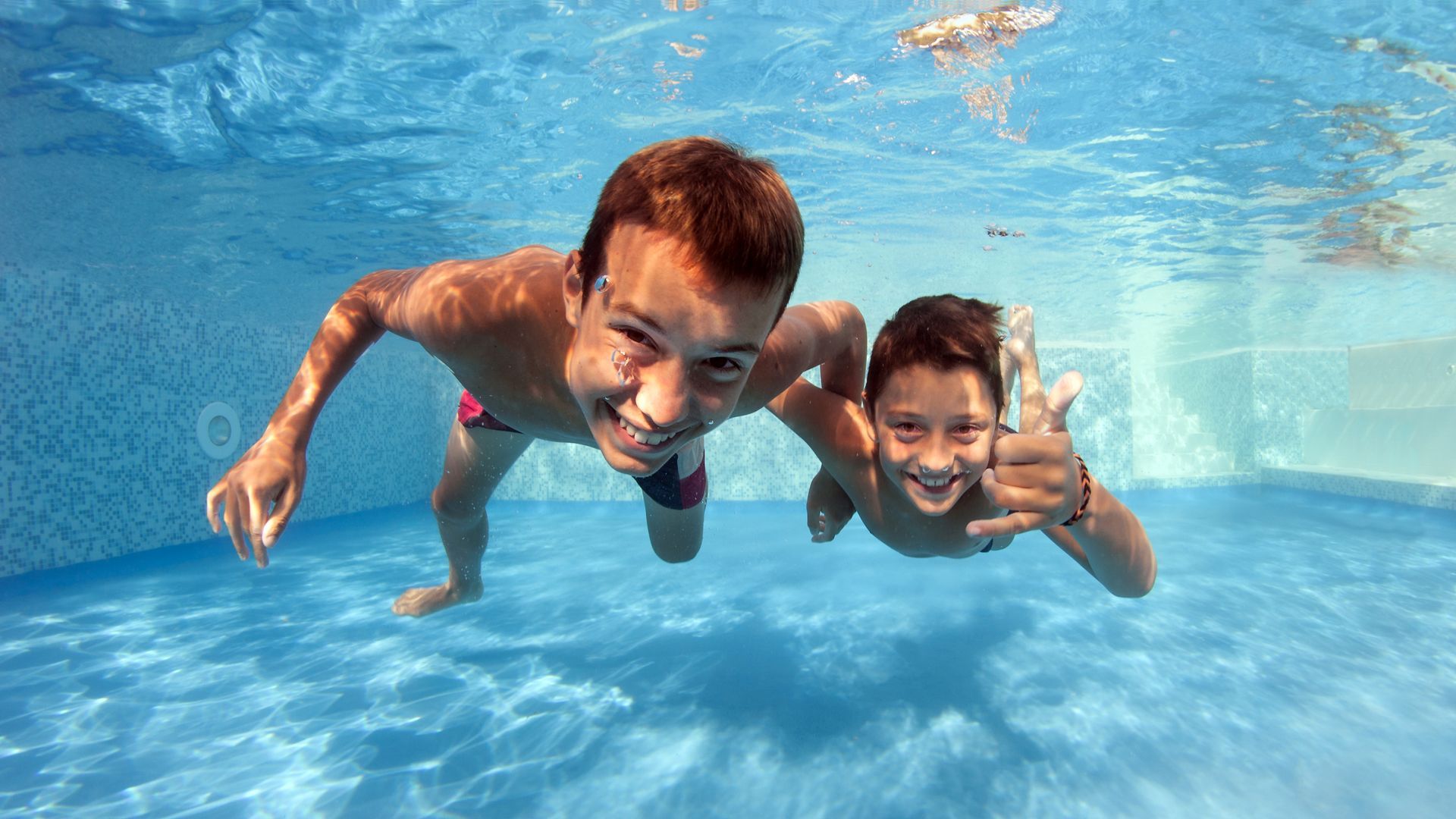 Two people smile while swimming underwater in a blue pool, one making a thumbs-up gesture.