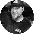 A man with a beard is wearing a hat and smiling in a black and white photo.