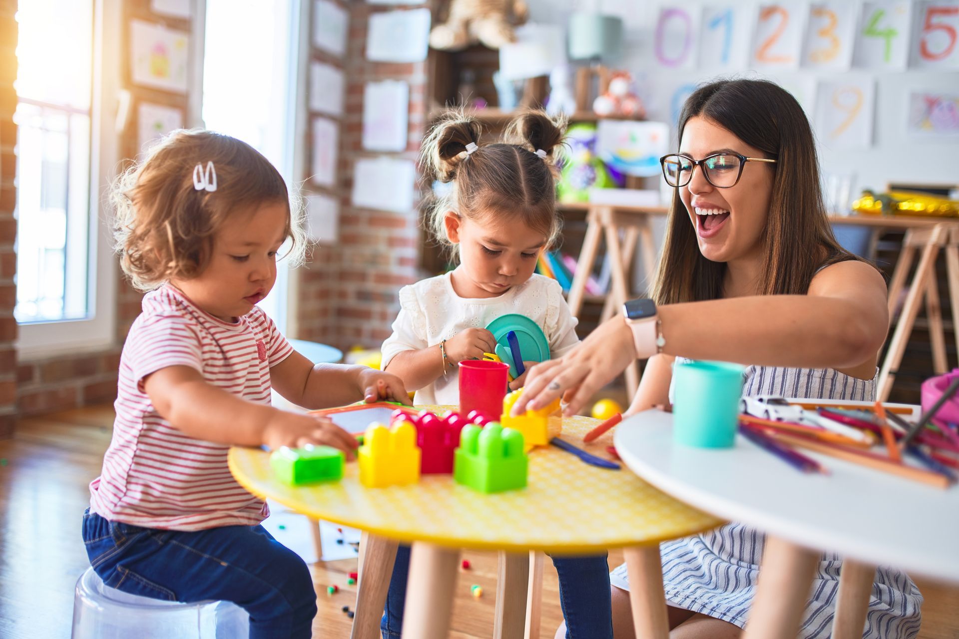 A woman and two toddlers playing with colorful blocks at a small table indoors.