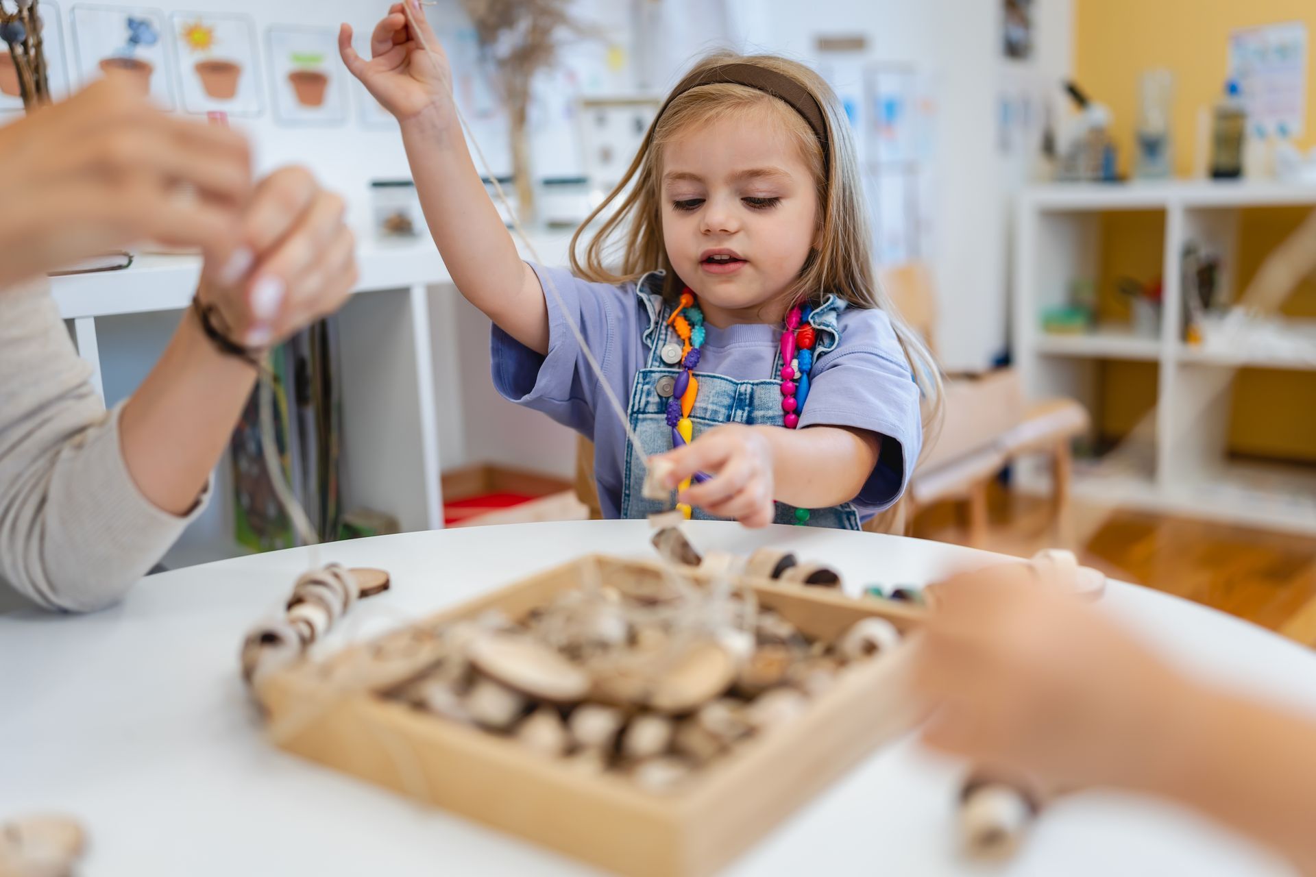 Young child stringing beads at a table with an adult in a classroom setting.