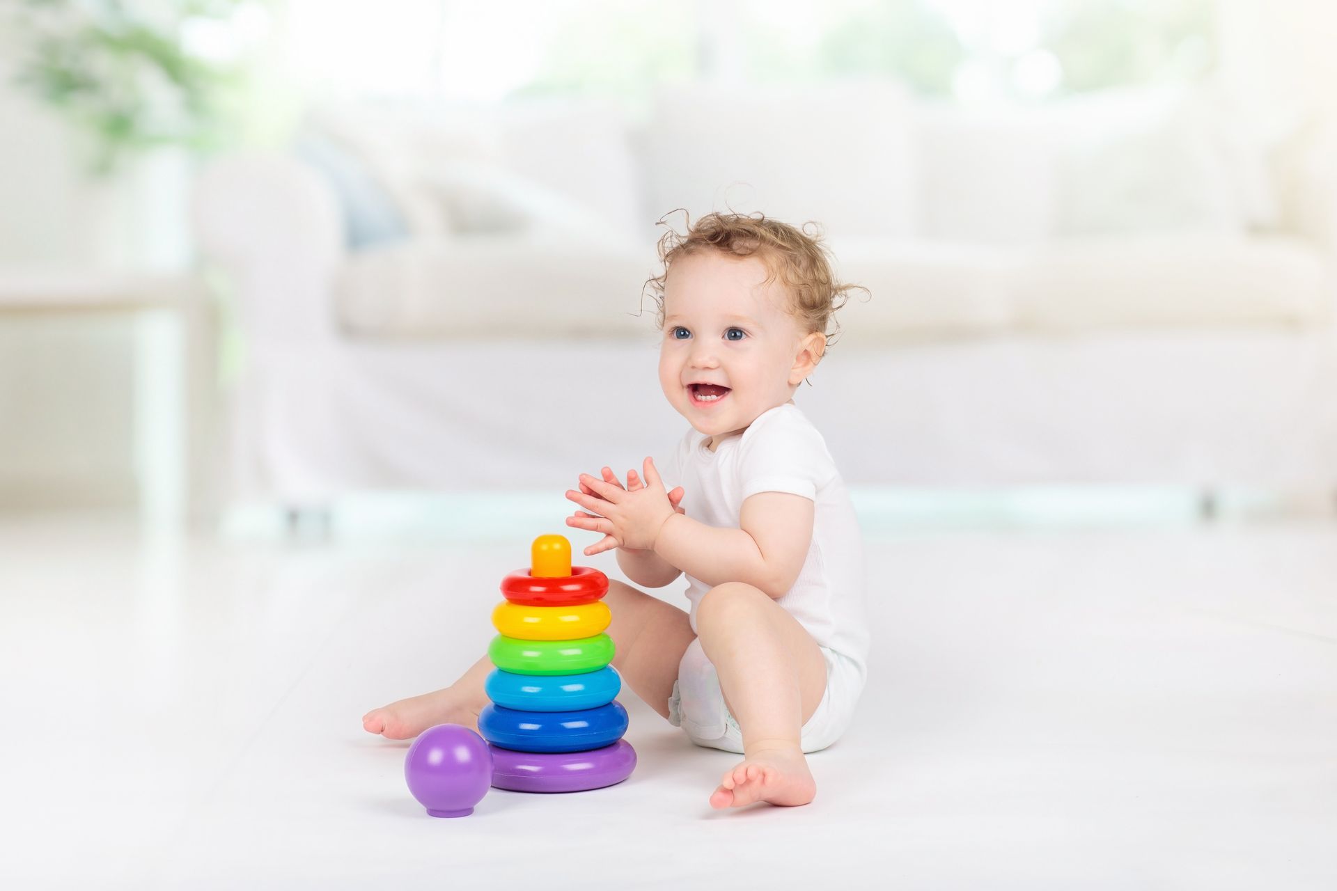 Happy baby playing with a colorful stacking toy on a white floor.