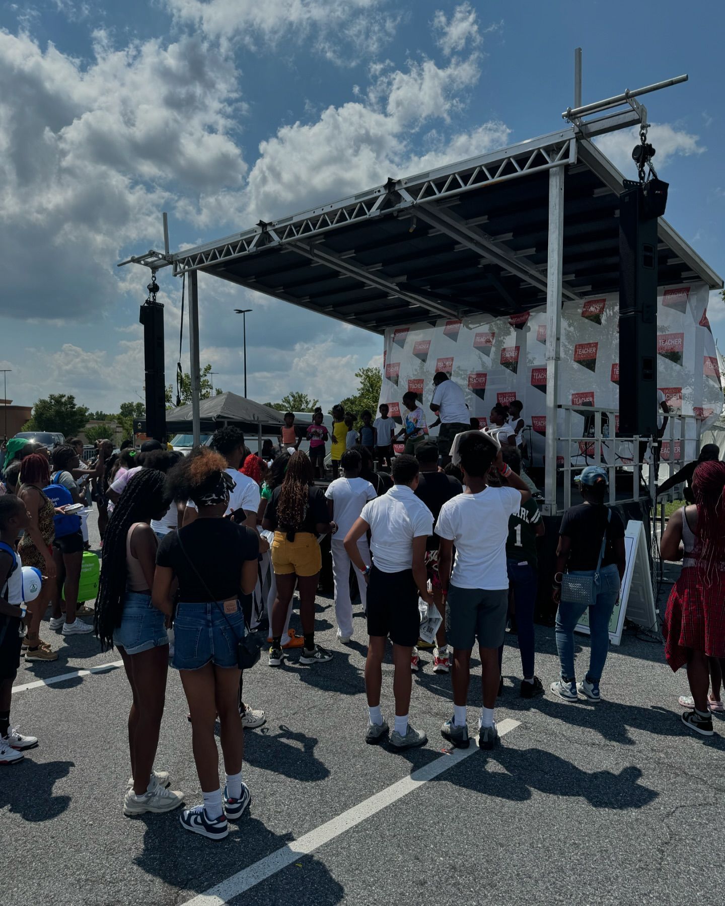 People gathered around an outdoor stage with sound equipment under a partly cloudy sky.