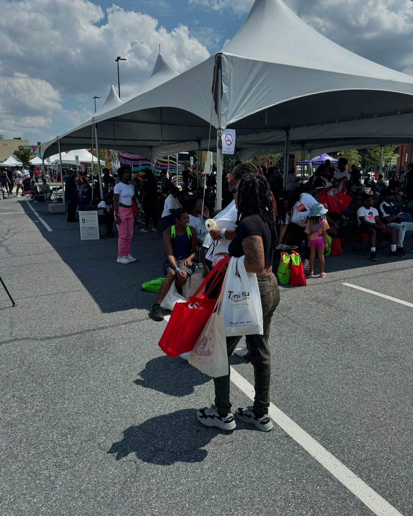 People at outdoor event under tents. Person holding shopping bags stands in front.