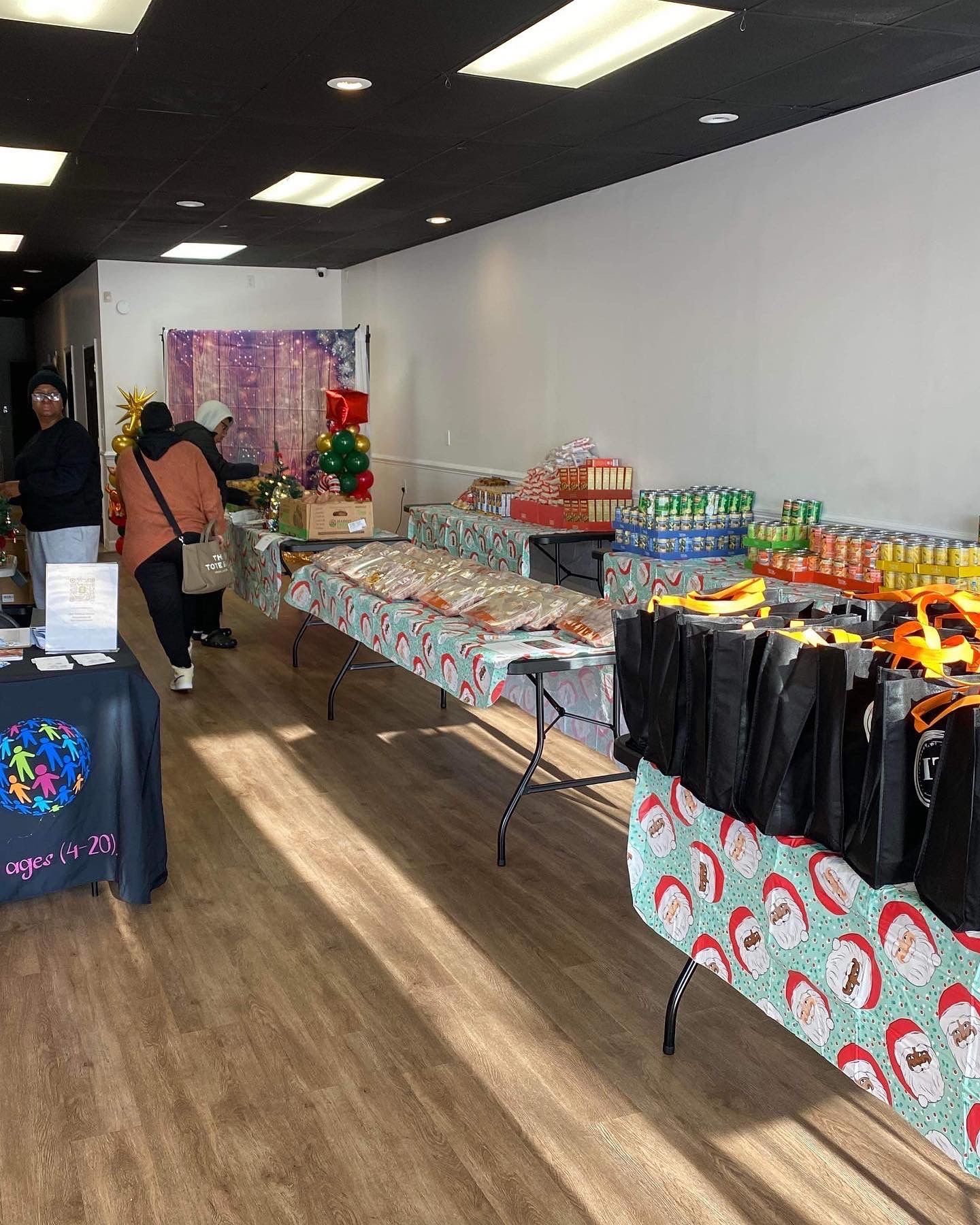 Store interior with tables of food and gifts. People browse items. Santa-themed tablecloths.