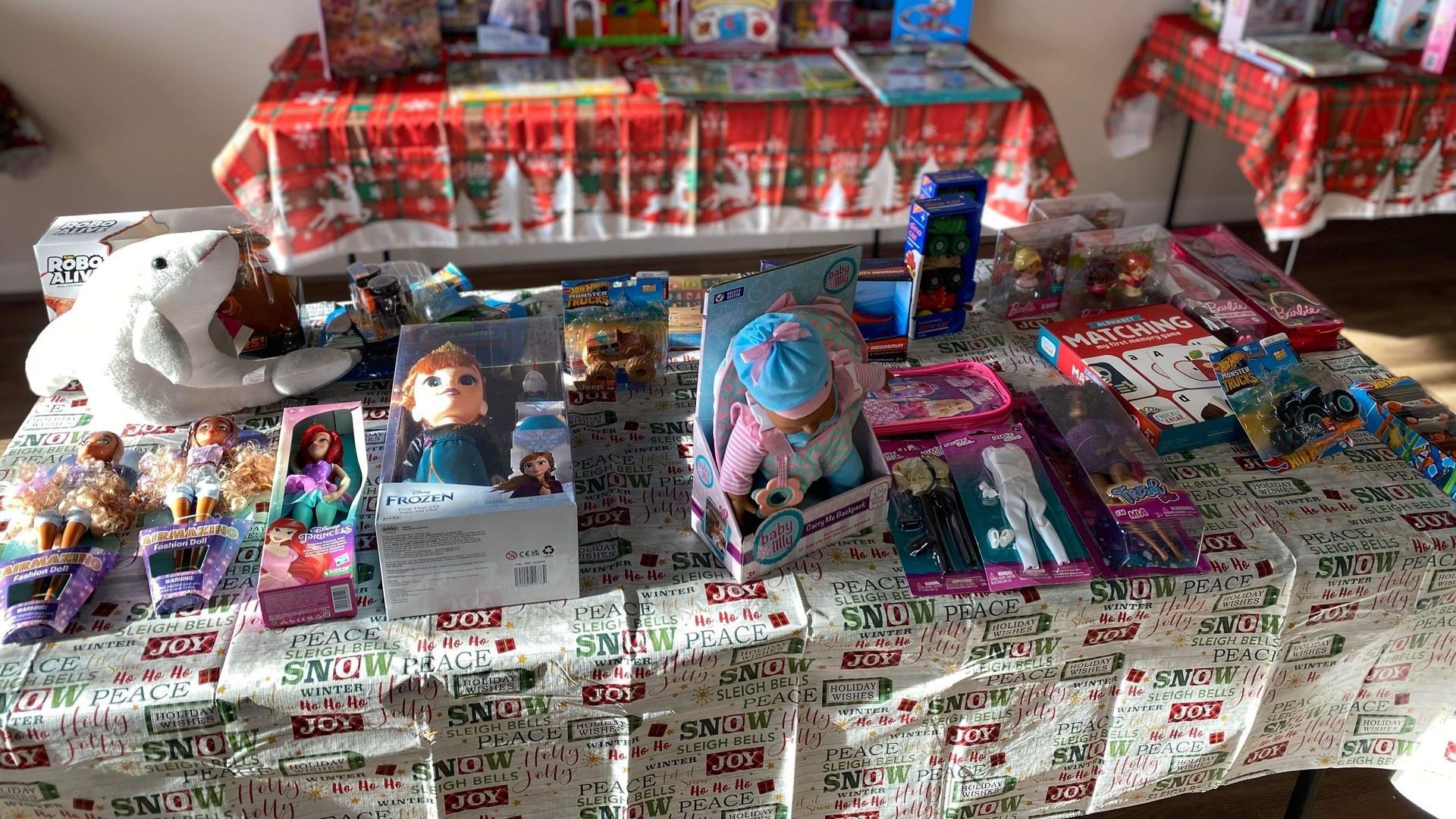 Toys on a table covered in a holiday-themed tablecloth.  Includes dolls, books, and other gifts.