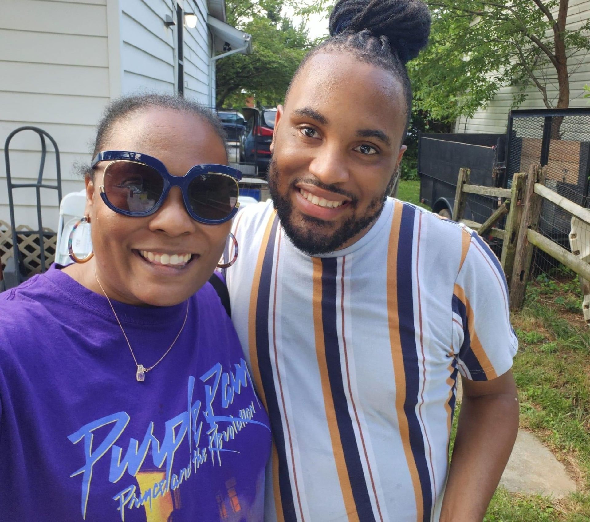 Two smiling people pose for a selfie outdoors. One wears a purple shirt, large sunglasses. The other has striped shirt, hair tied up.