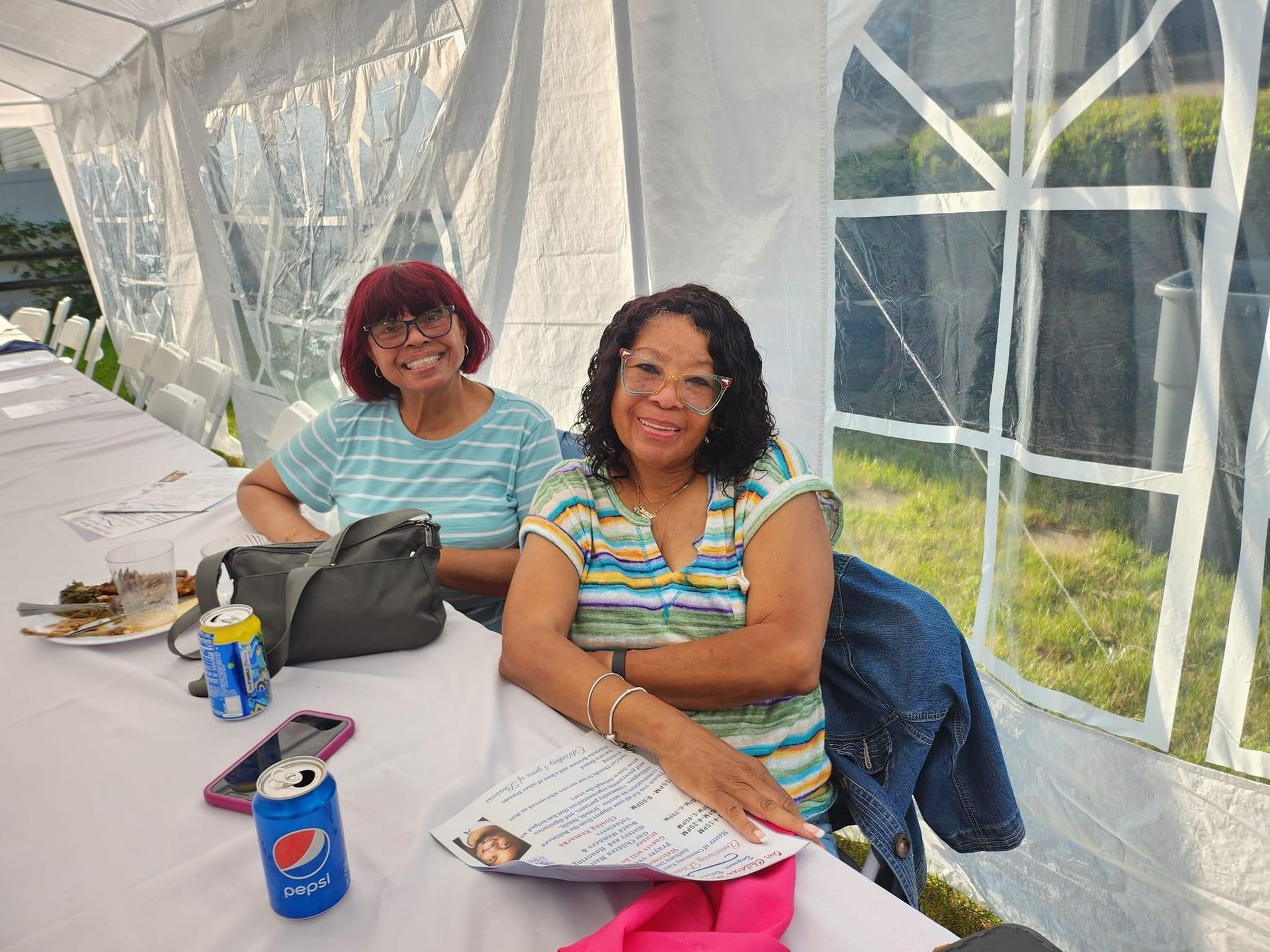 Two women smiling at a table in a tent. One with short red hair and a blue shirt, the other in a striped shirt.