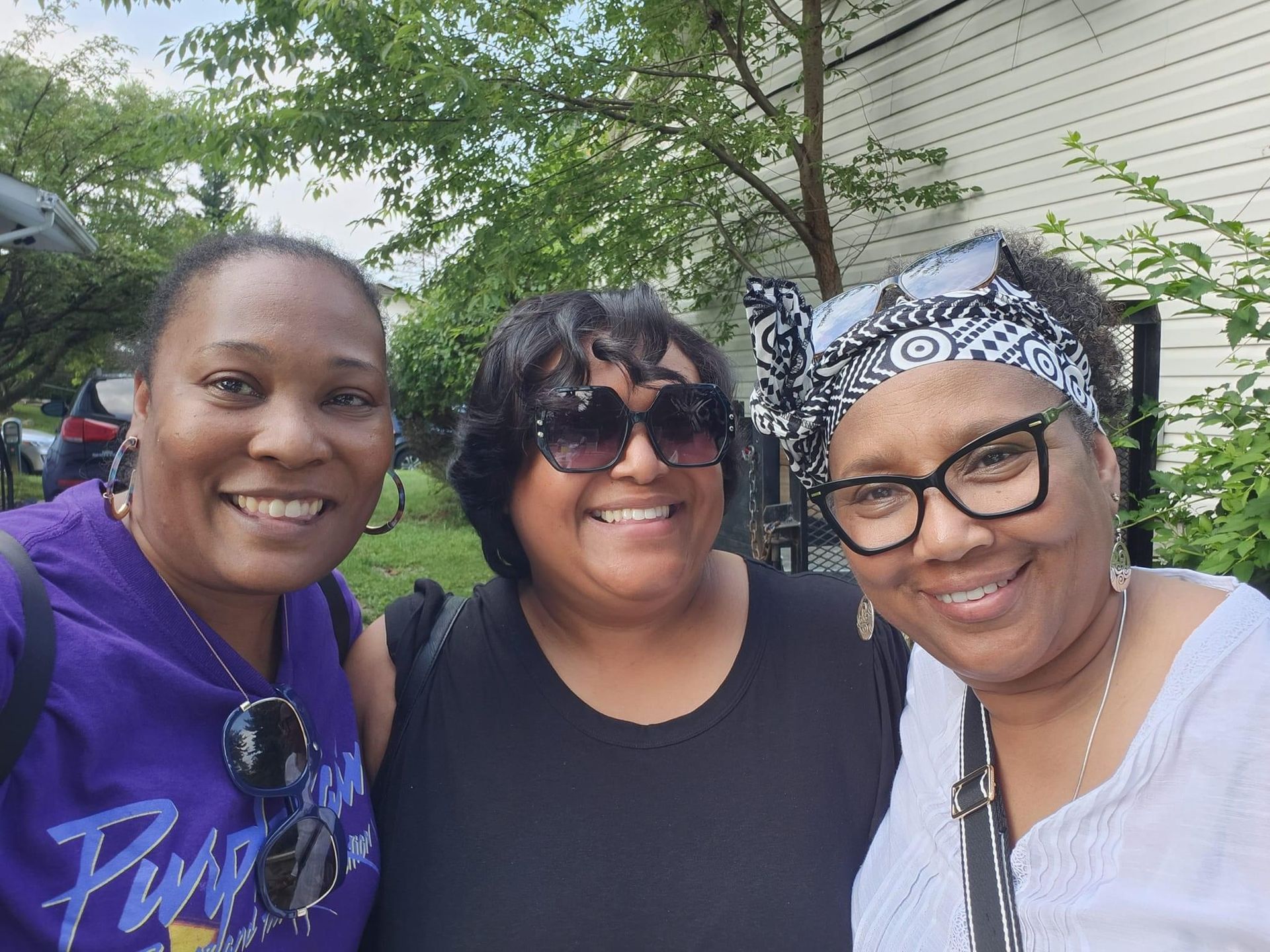 Three smiling people pose outdoors. One wears a purple shirt, another a black top, and the last a white shirt.