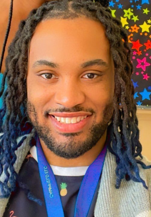 Man with dreadlocks, smiling, wearing a blue lanyard and gray sweater. Colorful star background.