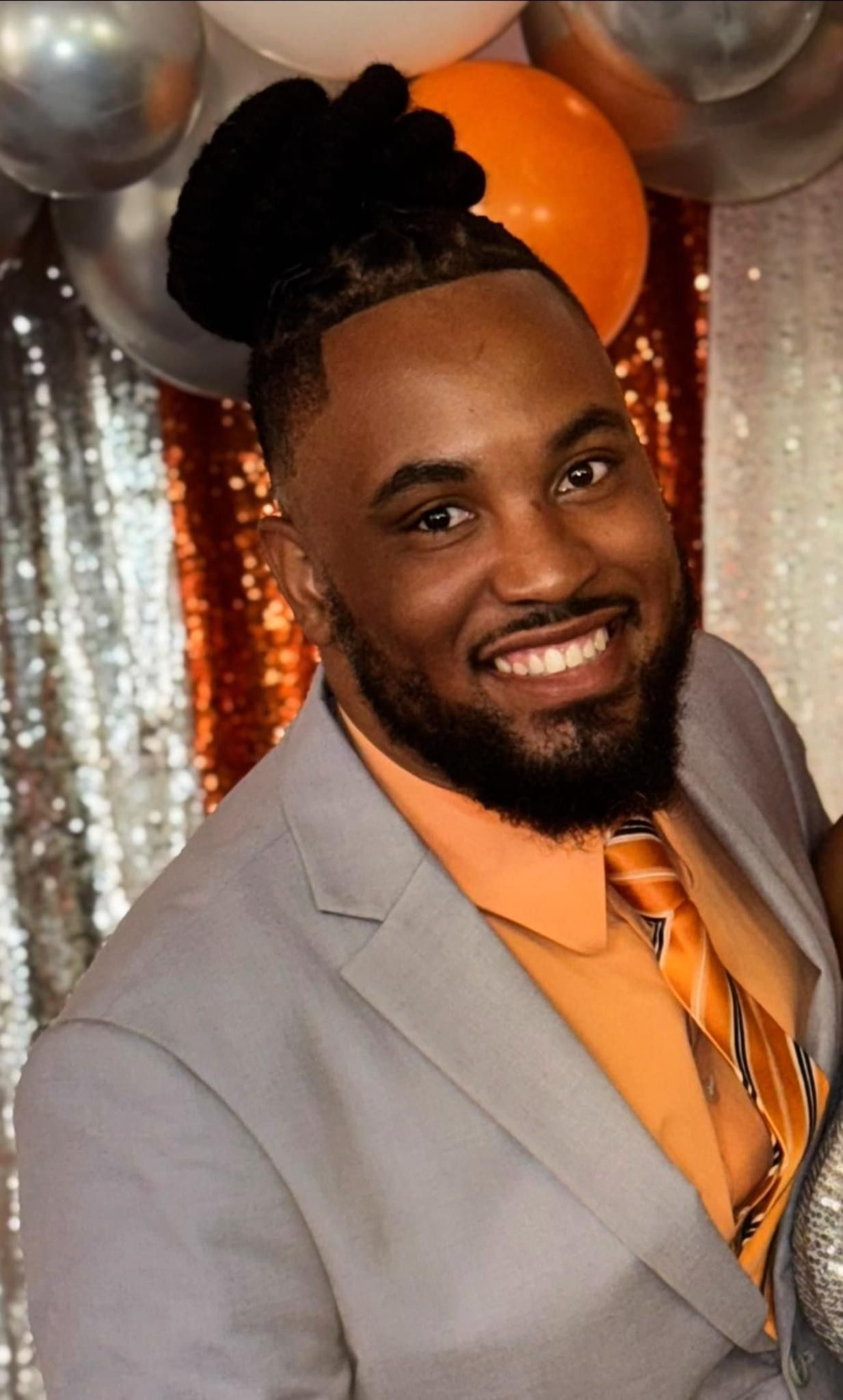 Man in gray suit smiles, orange shirt and tie. Black hair up, with balloons in background.