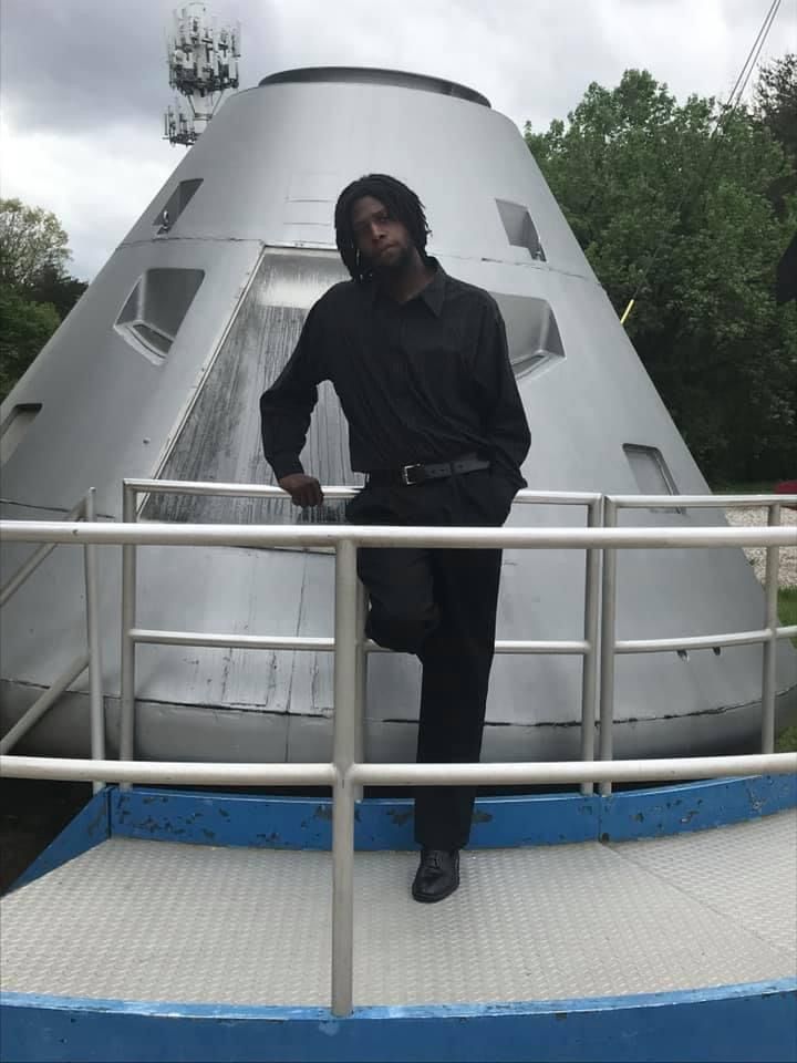 Person in black leans against a silver, cone-shaped structure with windows, outdoors on a cloudy day.