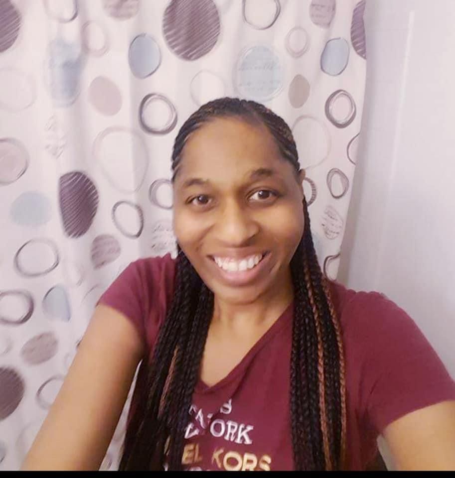 Woman with braided hair smiling, wearing a burgundy shirt, in front of a patterned shower curtain.
