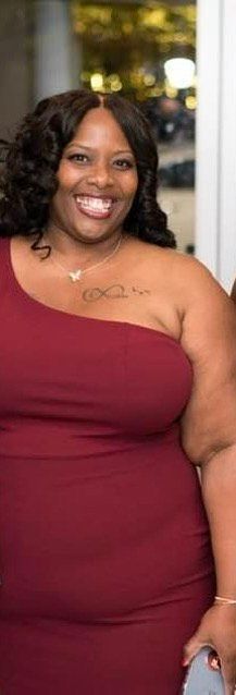 Woman in a maroon one-shoulder dress smiles at the camera, wearing a necklace and holding a phone.
