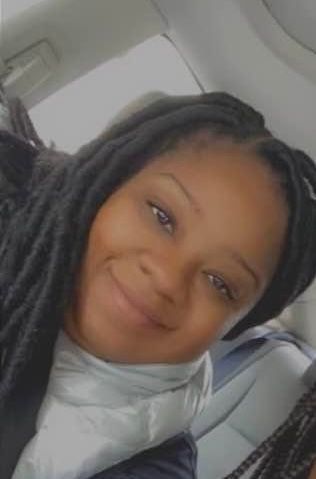 Woman with dark locs smiles, wearing a light colored scarf, possibly in a vehicle.