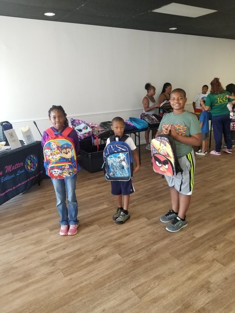 Three children smiling, holding backpacks indoors. A table with supplies is visible. People are in the background.