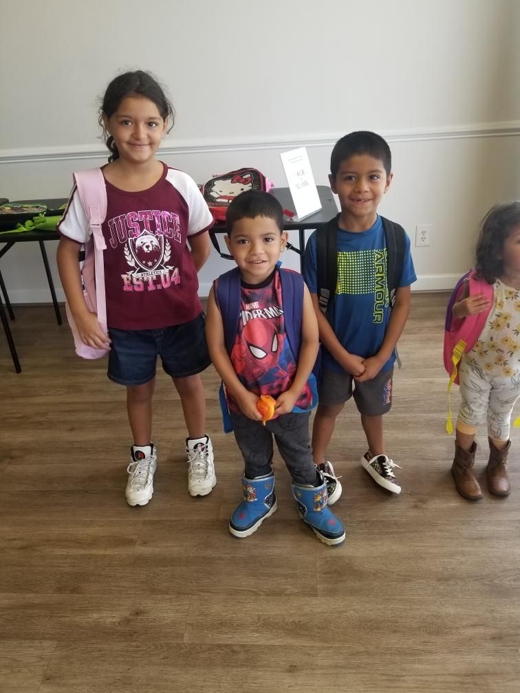 Four children with backpacks, standing on a wood floor. Smiling, wearing various outfits.