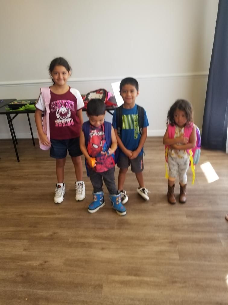 Four children standing indoors, wearing backpacks. Two children are looking at the camera. Wooden floor, neutral walls.
