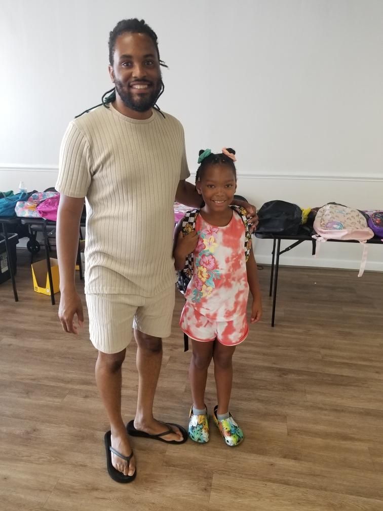 Man and child pose. Both smile. Backpacks and tables in the background. Light-colored room.