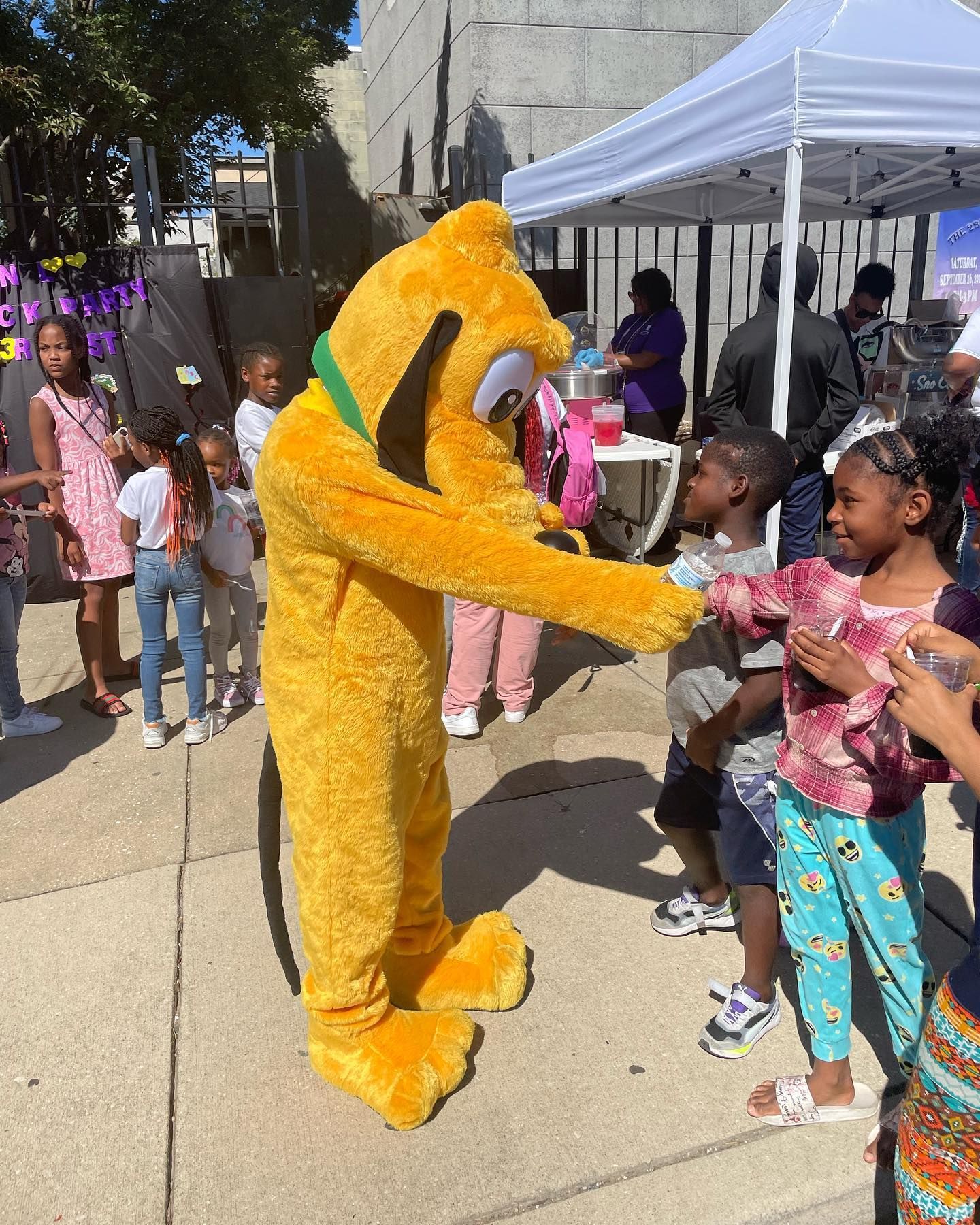 Pluto character interacting with children at an outdoor event, some children clapping and smiling.