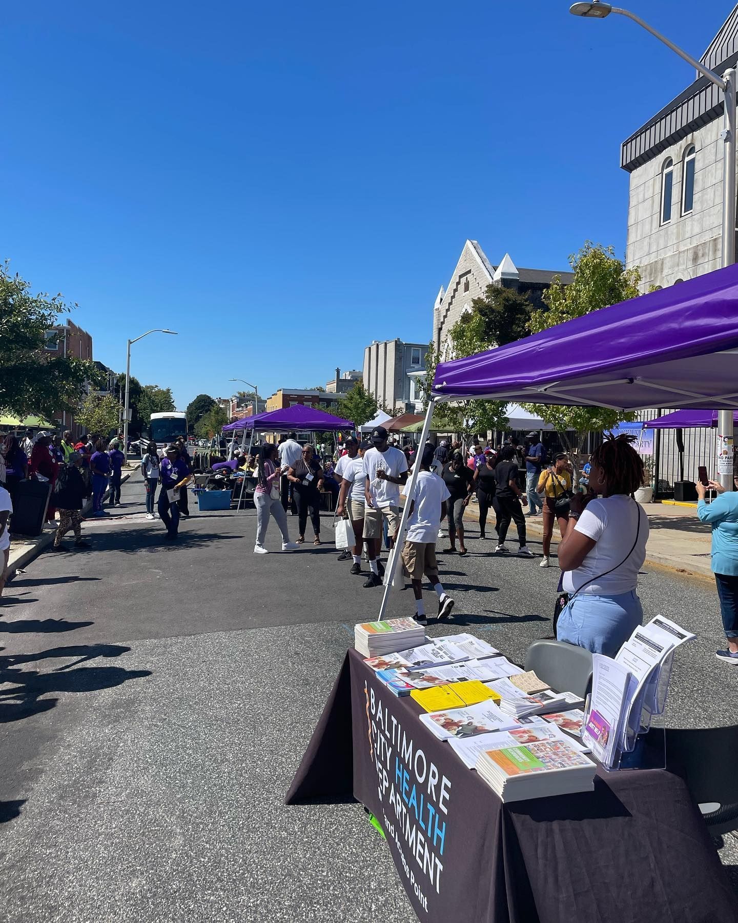 Street fair with purple tents and people, under a sunny sky. A table with pamphlets.