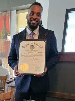 Man in a blue suit holds a Maryland State proclamation, smiling. Indoor restaurant setting.