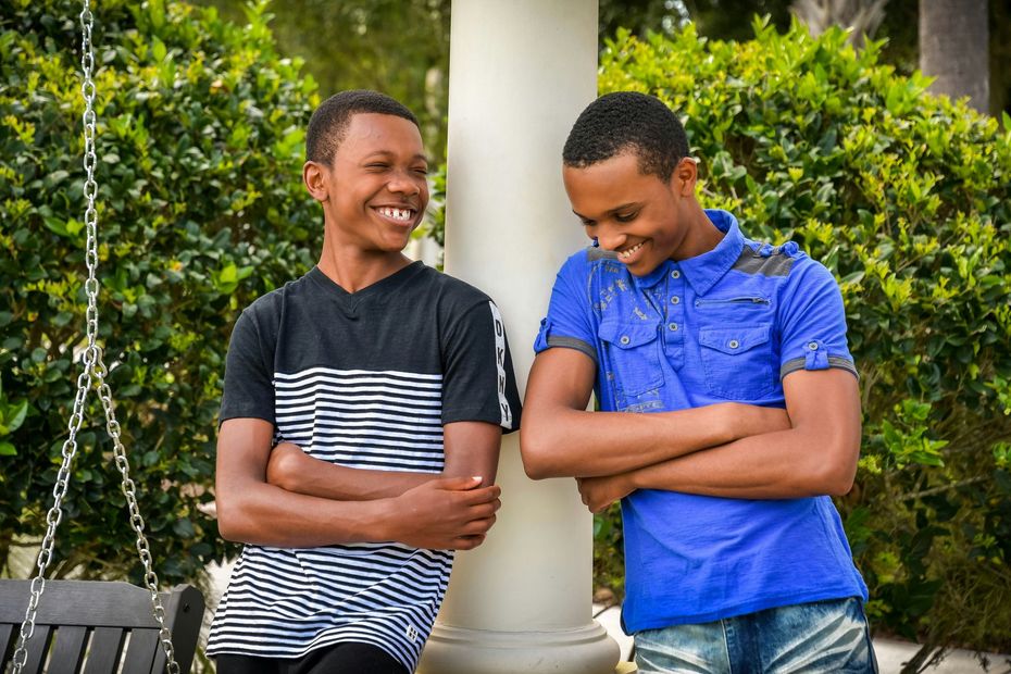 Two young people smiling, leaning against a white pillar outside. One wears a striped shirt, the other a blue shirt.