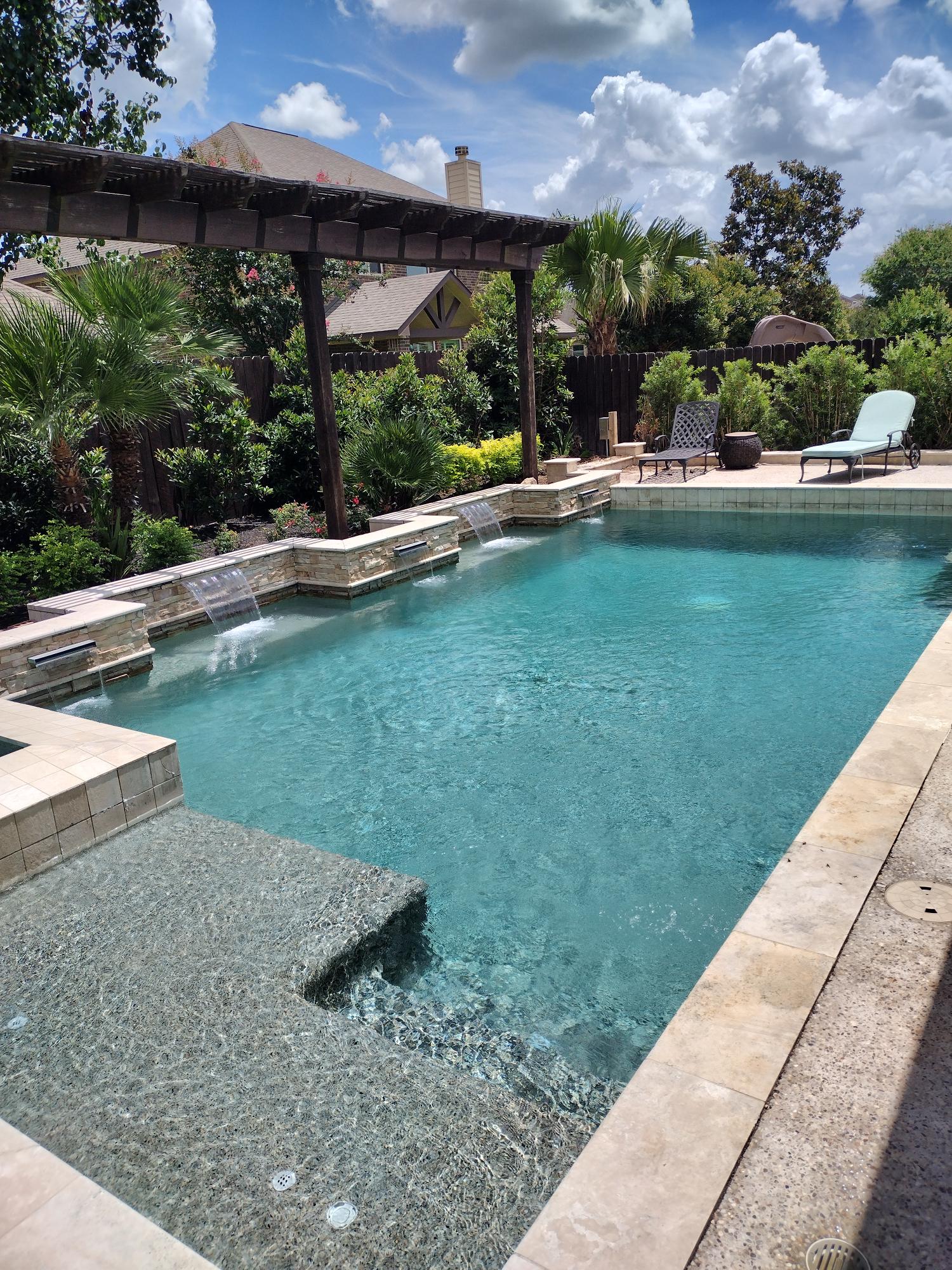 Backyard swimming pool with clear blue water, stone coping, pergola, and lounge chairs under a partly cloudy sky