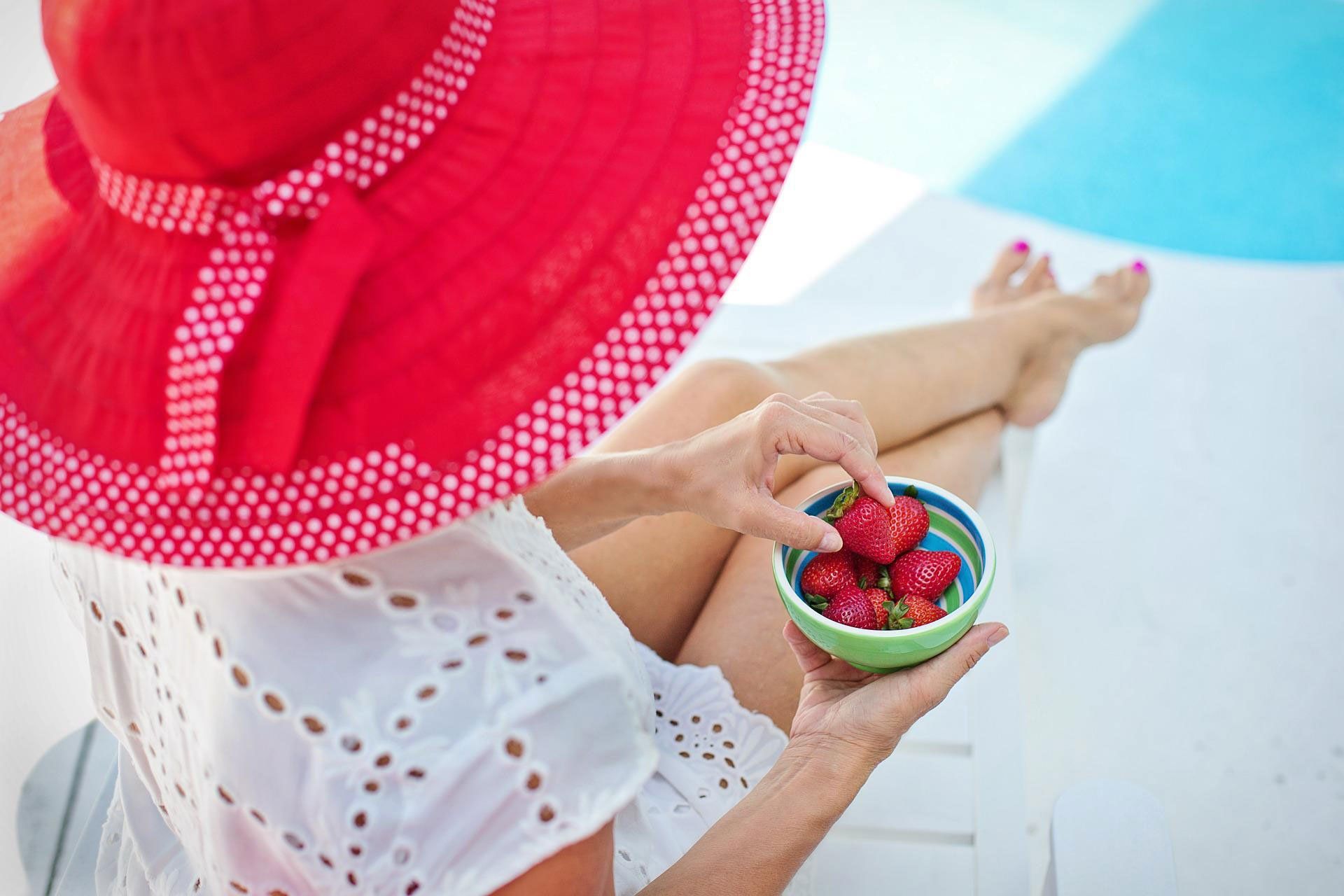 Person in a red sunhat holding a bowl of strawberries by a pool