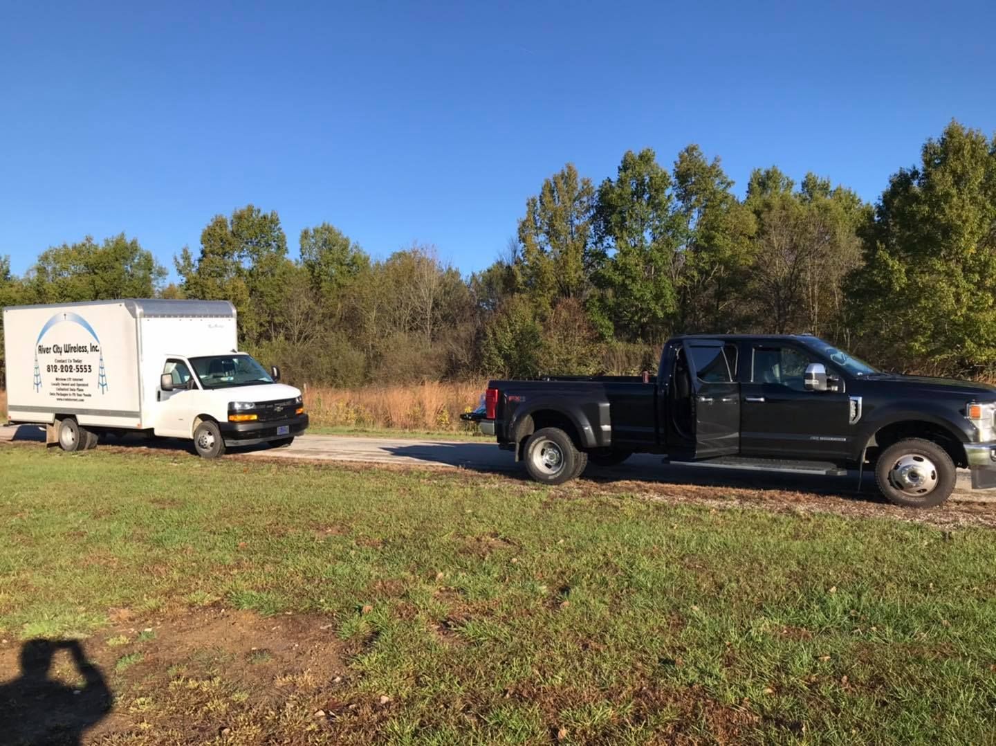 A black pickup truck towing a white box truck on a dirt road in a field on a sunny day.