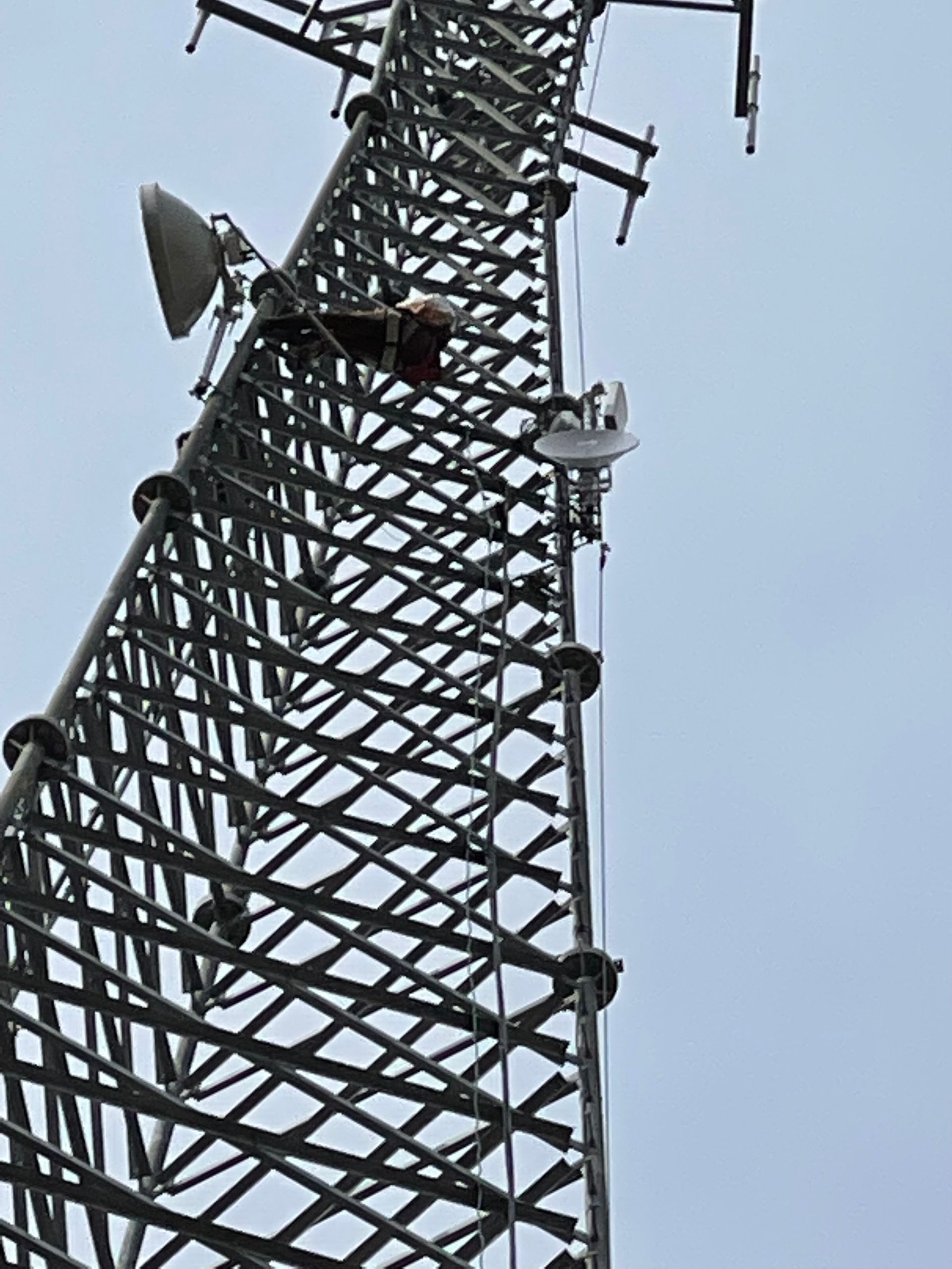 Person on a communications tower, working with equipment. Metal structure against a cloudy sky.