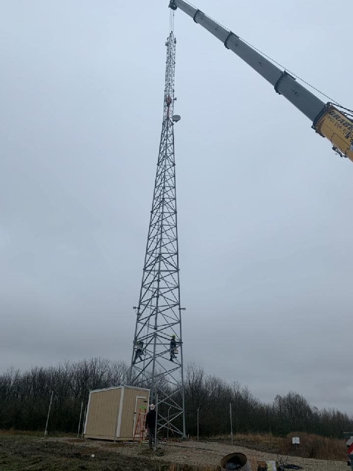 Crane lifting a metal telecommunications tower; workers on the structure, with equipment and a small building at the base, under a cloudy sky.