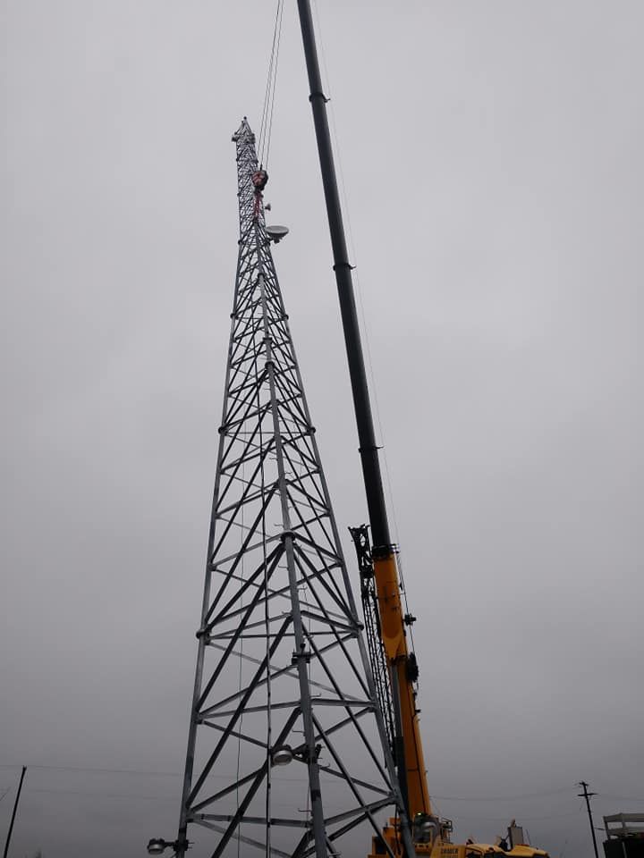 Crane lifting section of a metal communication tower under construction on a cloudy day.
