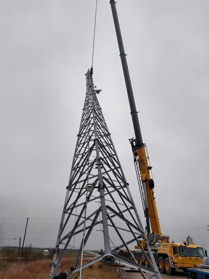 A crane lifting sections of a metallic telecommunications tower under a cloudy sky.