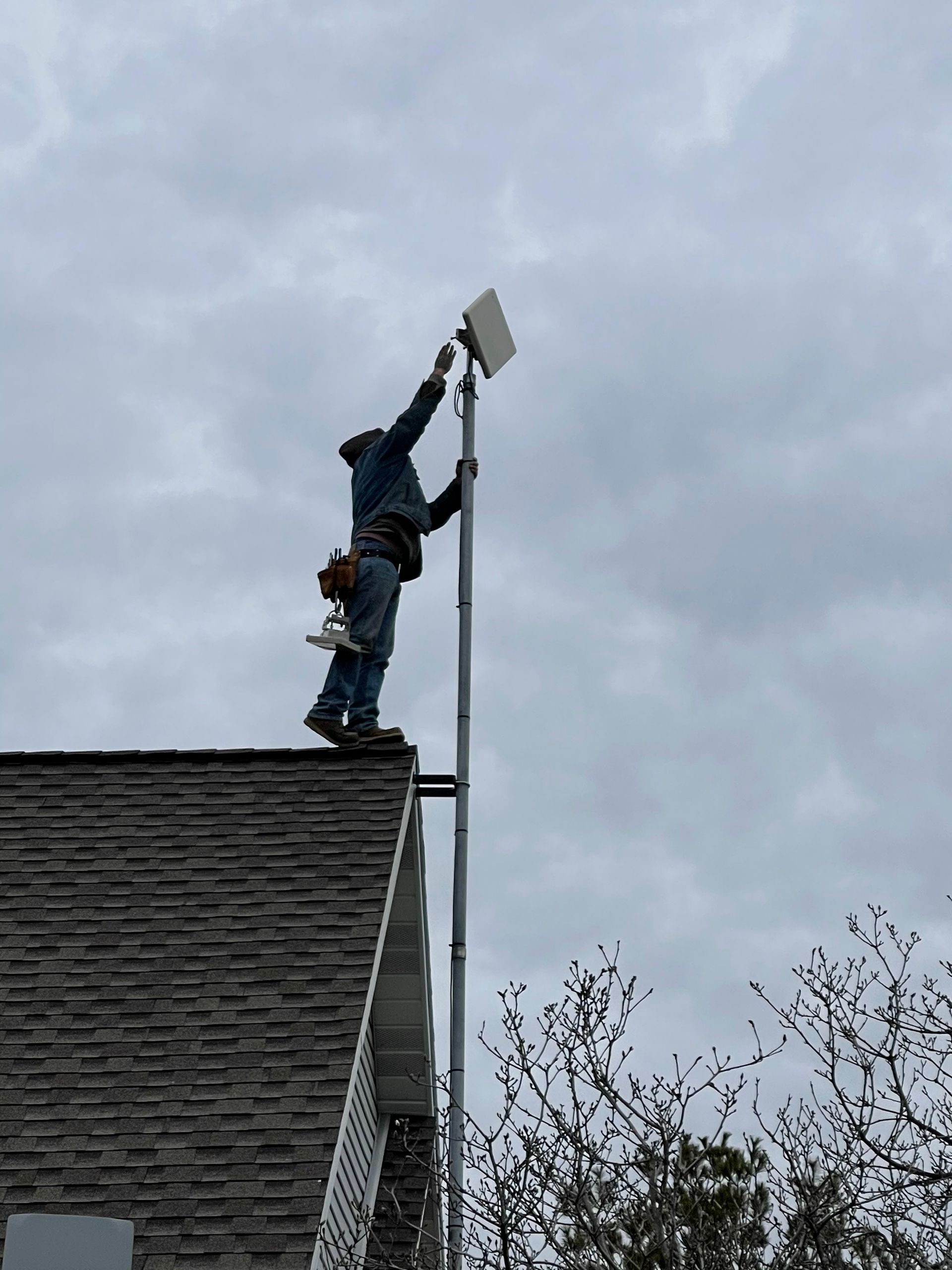 Person on a rooftop installing a white rectangular antenna on a pole, against a cloudy sky.