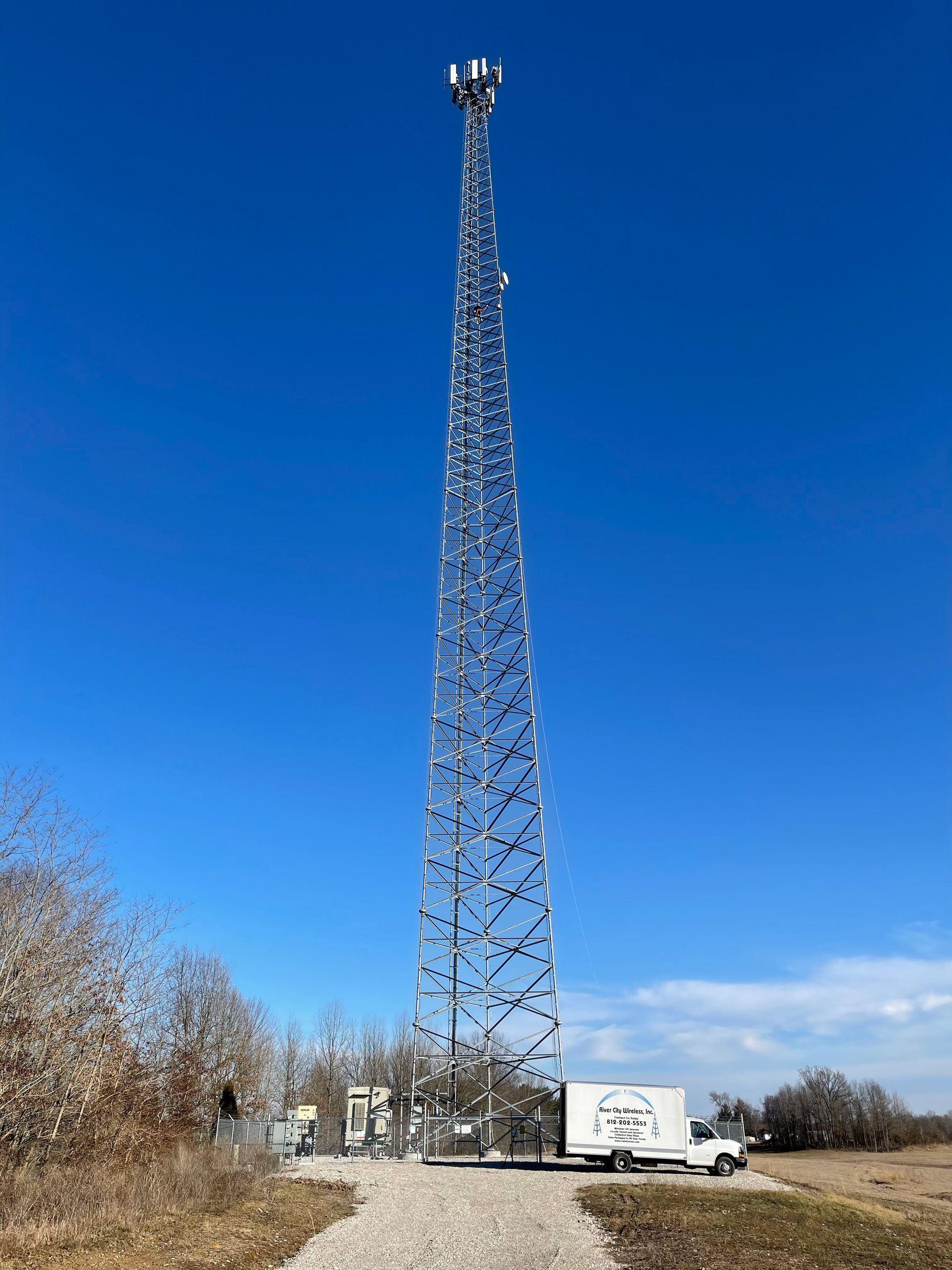 Tall metal cell tower on a sunny day with a service truck parked below.