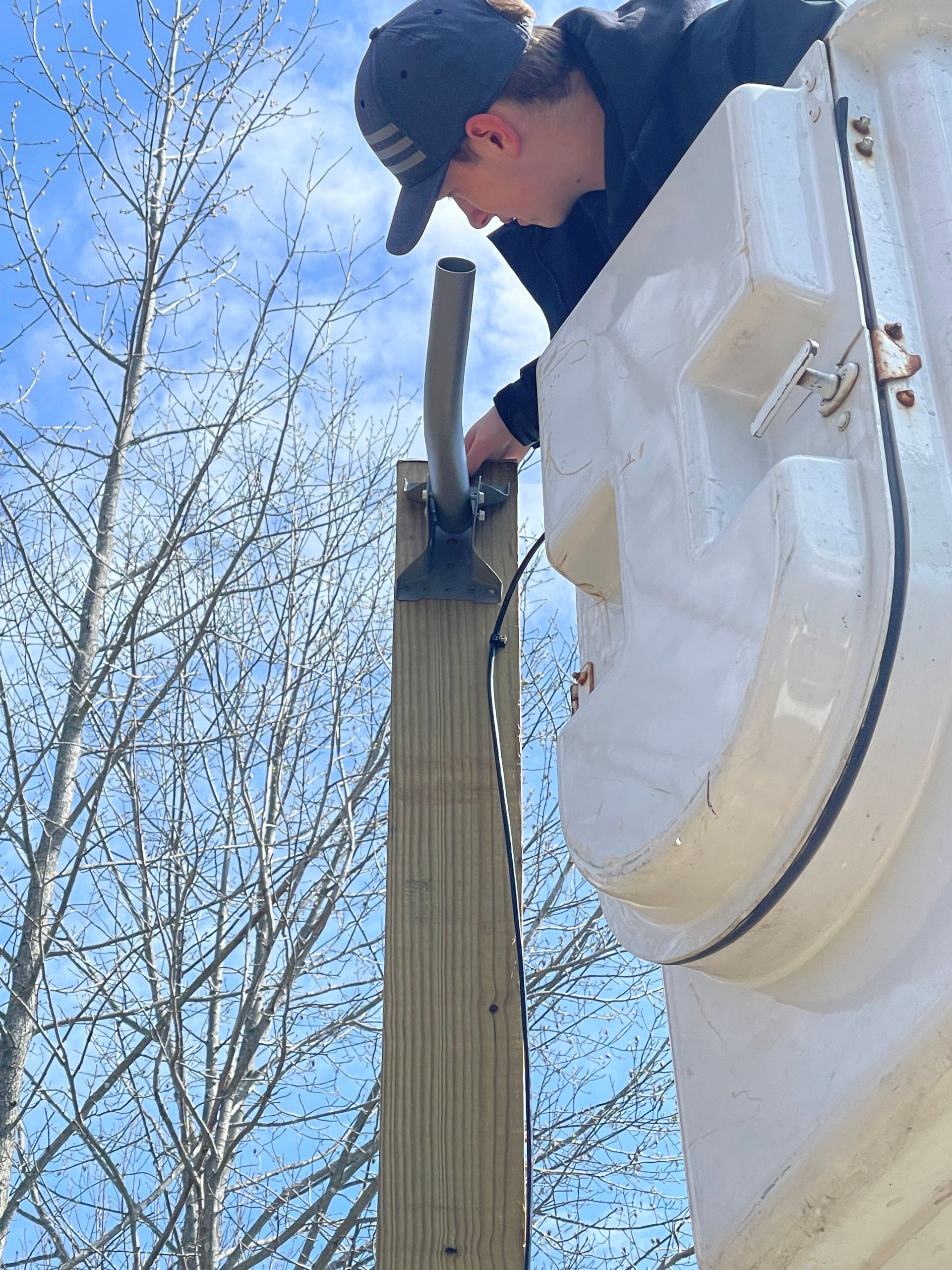 Person in a lift bucket installing a metal arm onto a wooden post outdoors.