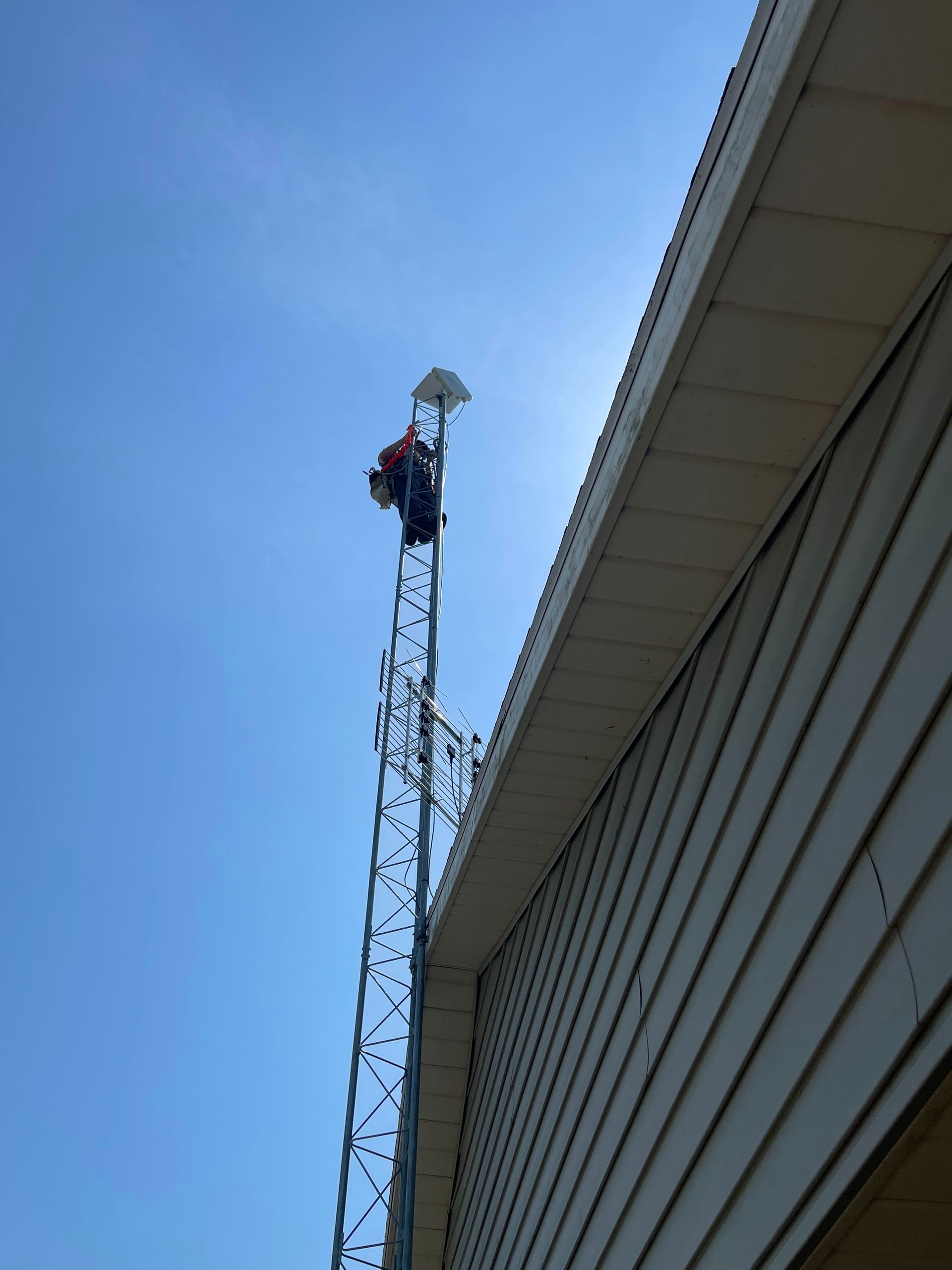 Person on a tall metal tower, likely installing equipment, next to a building under a blue sky.