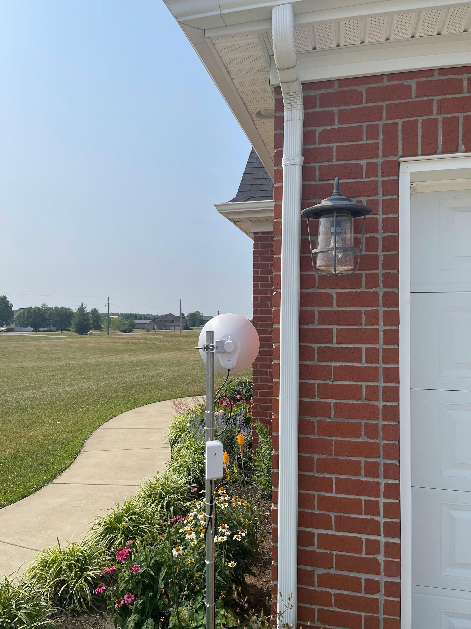 A white sphere-shaped weather device mounted next to a red brick building, near a driveway and grassy area on a sunny day.