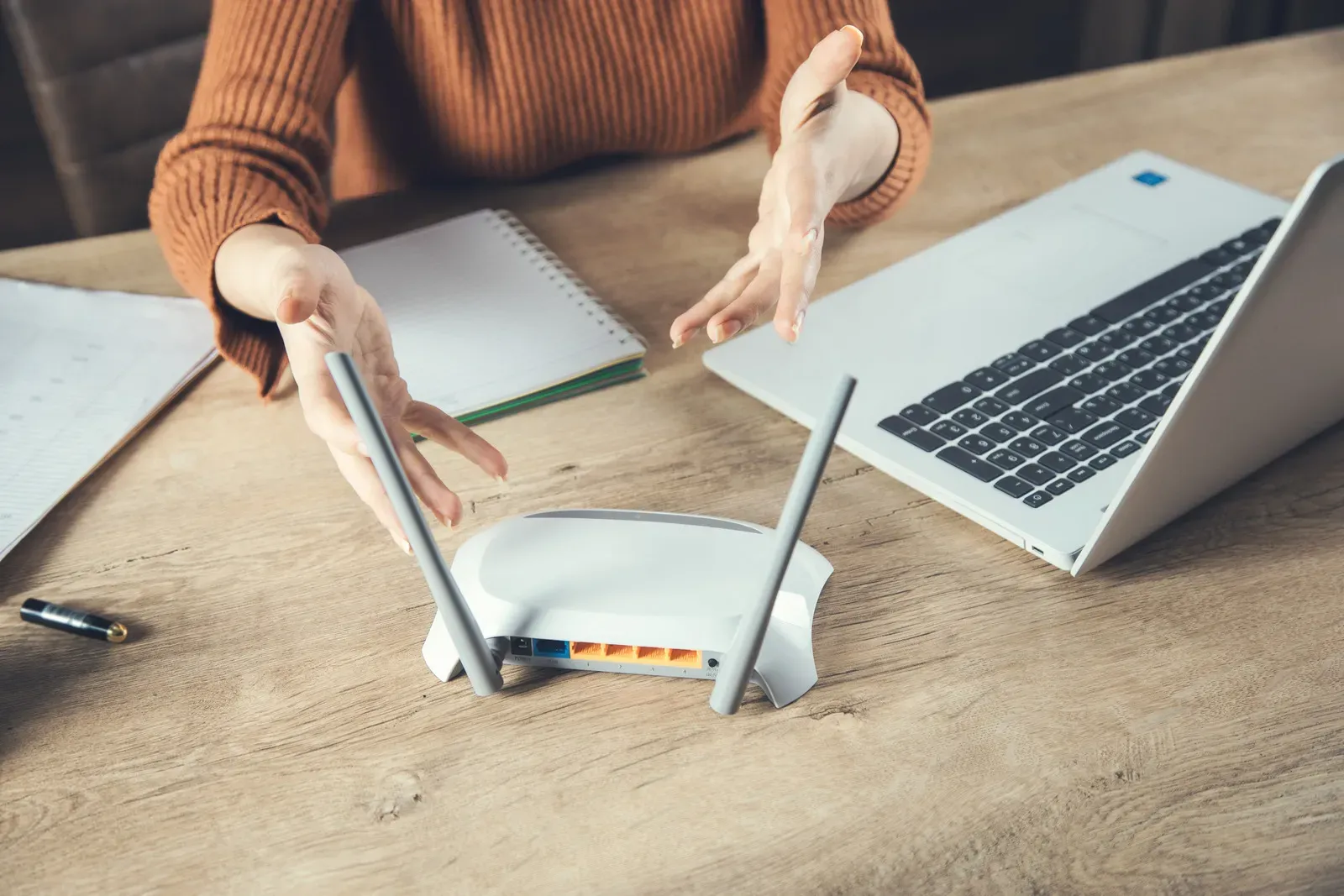 Person frustrated, gesturing at a router on a desk with a laptop and notebooks.