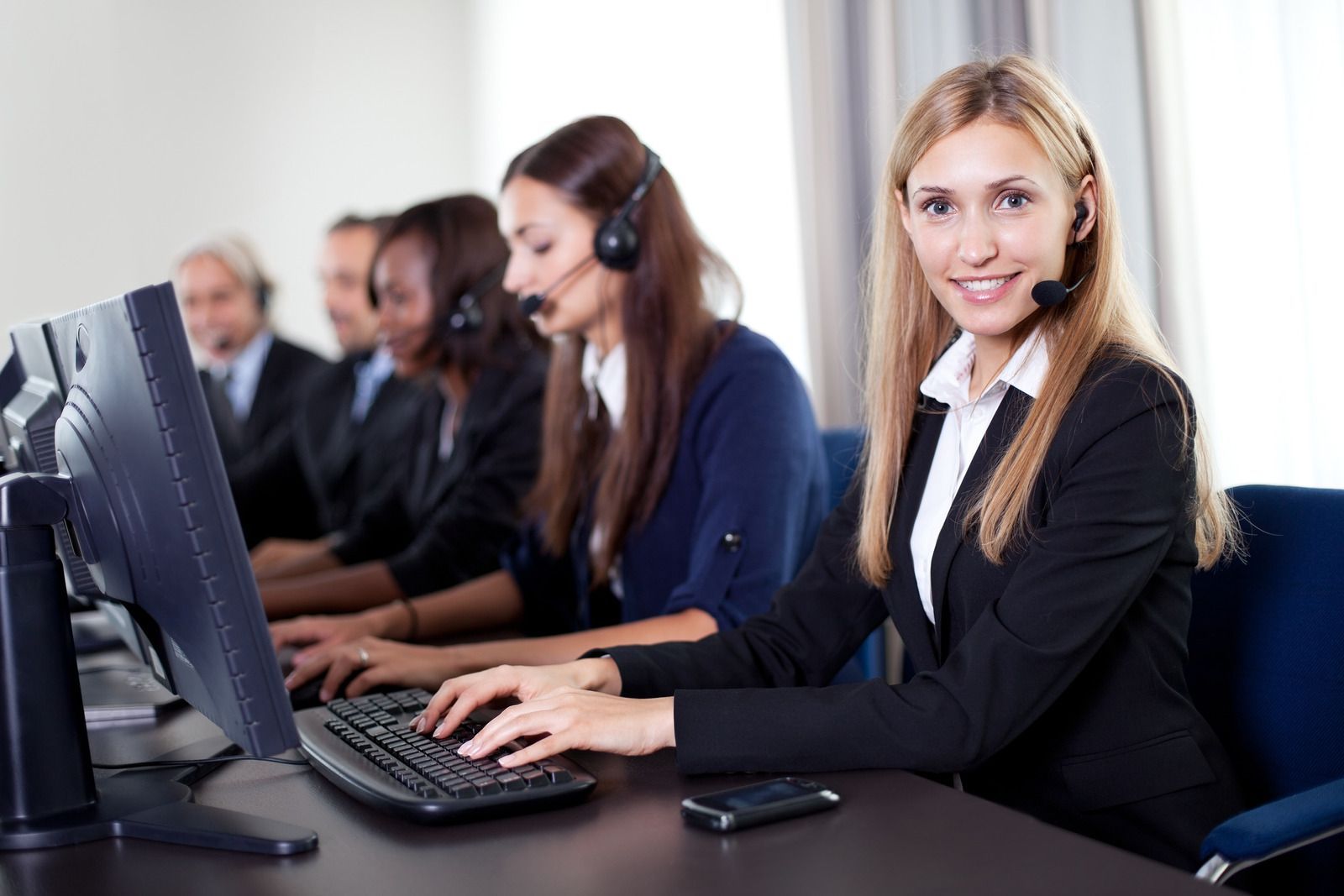 People in business attire working at computers with headsets in a call center setting.